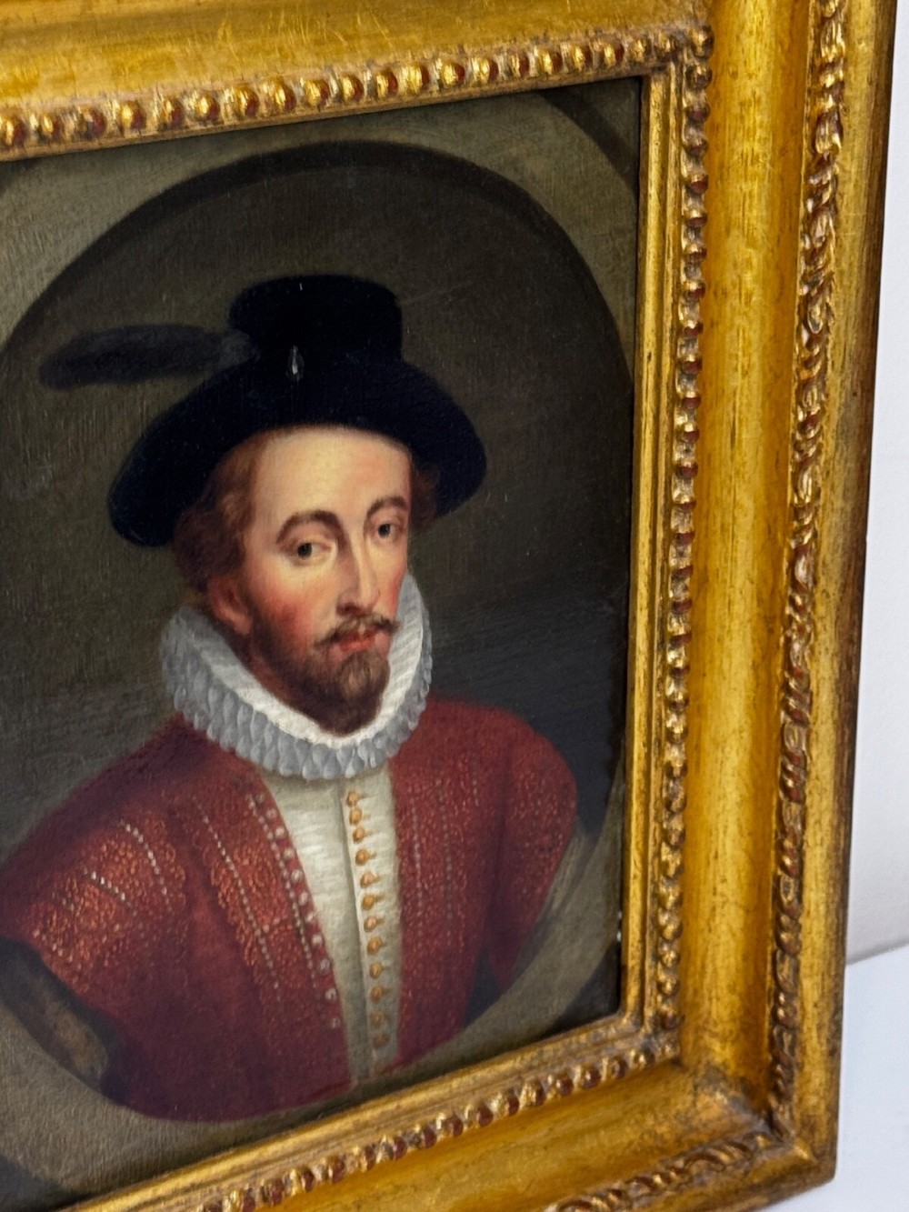 An 18th-century oil portrait of a man in a black hat, ruff collar, and crimson doublet set inside a carved gold-gilded frame. View XV