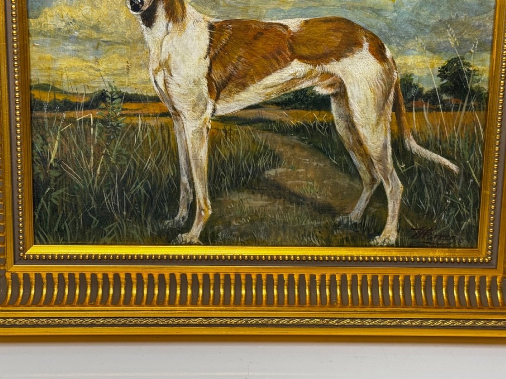 An antique Edwardian oil painting depicting a tan and white lurcher dog in a landscape, set within an ornate gilded frame. View X
