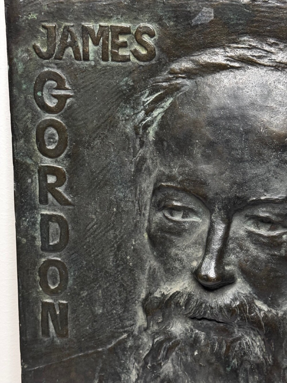 A rectangular dark bronze bas-relief plaque displaying a portrait of a bearded man with raised lettering and an incised signature. View VI