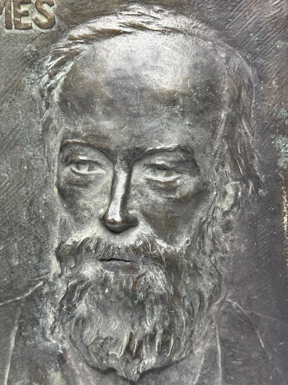 A rectangular dark bronze bas-relief plaque displaying a portrait of a bearded man with raised lettering and an incised signature. View VII