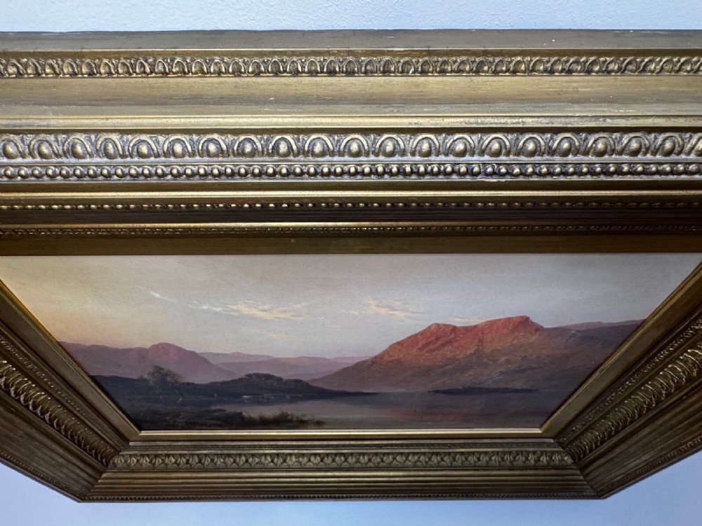 An antique oil painting depicting a lake surrounded by mountains at sunset, enclosed within a carved gold frame. View X