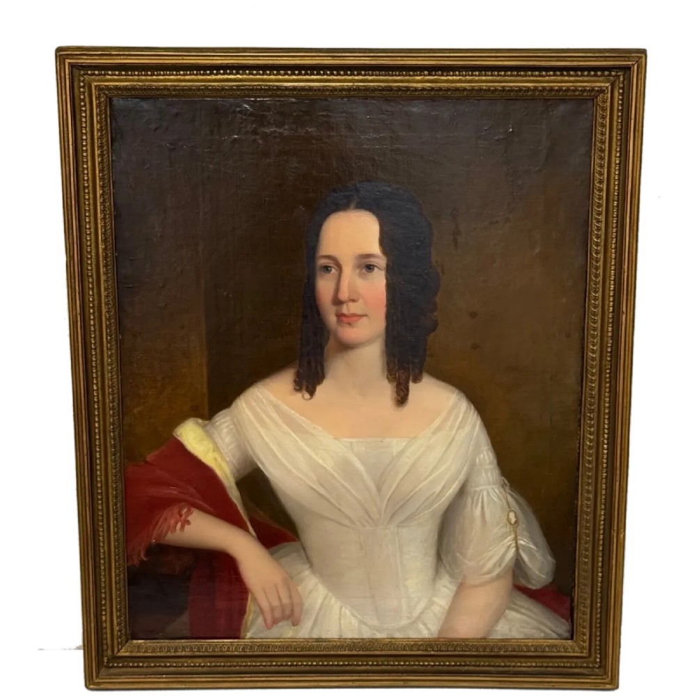 An antique oil painting depicting a woman in a white gown and red shawl set within a beaded giltwood frame.