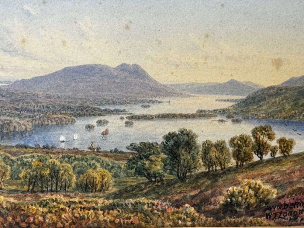 An antique Edwardian watercolour painting depicting Lake Windermere with distant hills, trees, and small boats, framed in gilt with a cream mount. View X