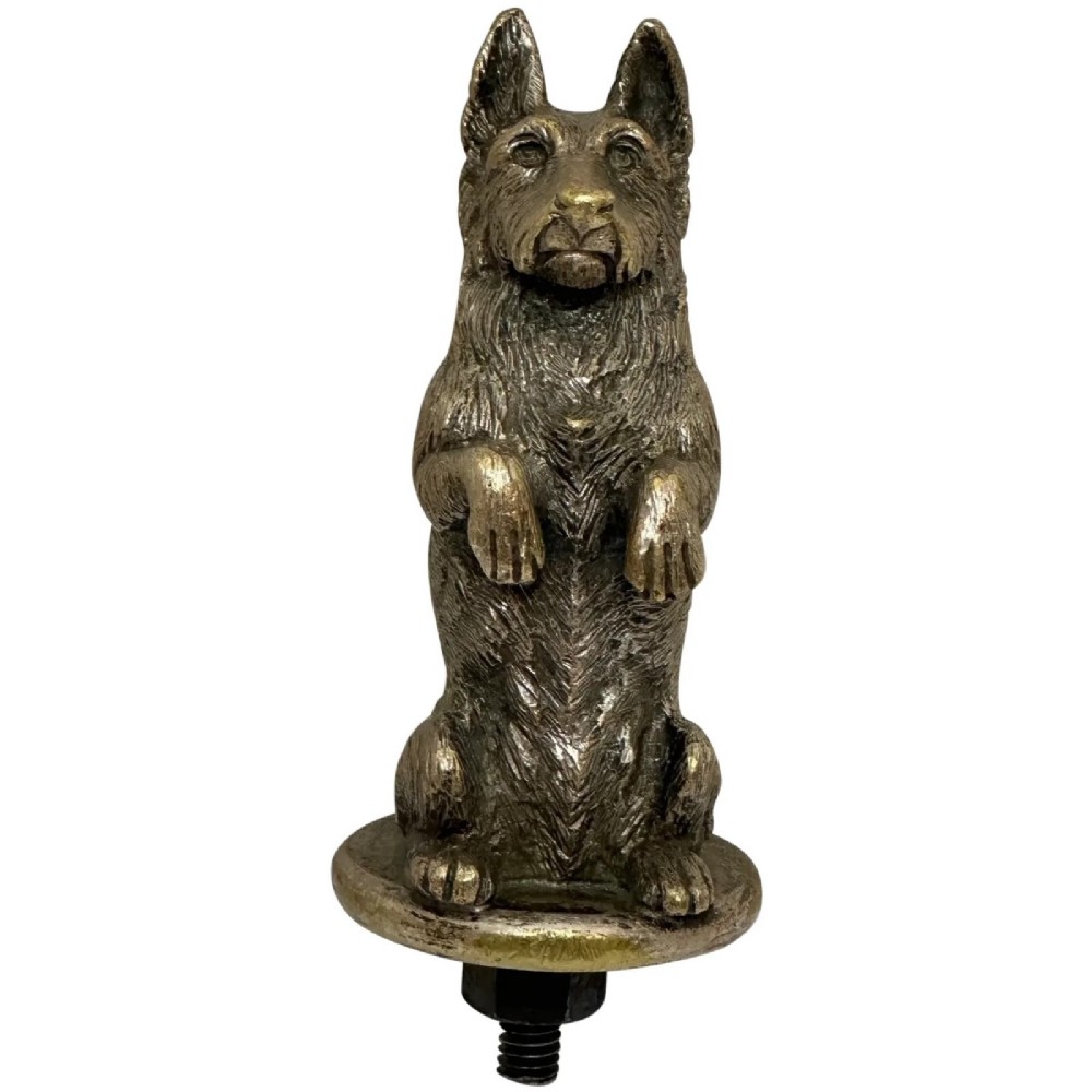 A cast bronze car mascot depicting a Scottish Terrier sitting in a begging pose mounted on a small circular base with a threaded bolt.