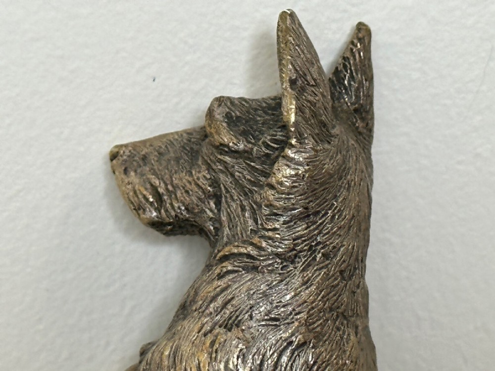 A cast bronze car mascot depicting a Scottish Terrier sitting in a begging pose mounted on a small circular base with a threaded bolt. View III