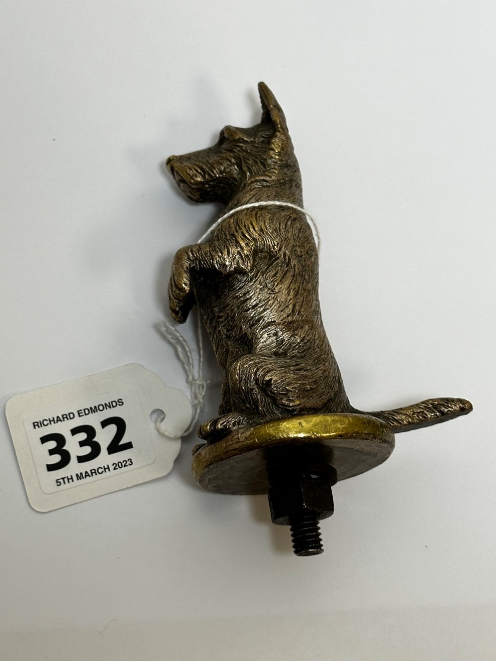 A cast bronze car mascot depicting a Scottish Terrier sitting in a begging pose mounted on a small circular base with a threaded bolt. View IX