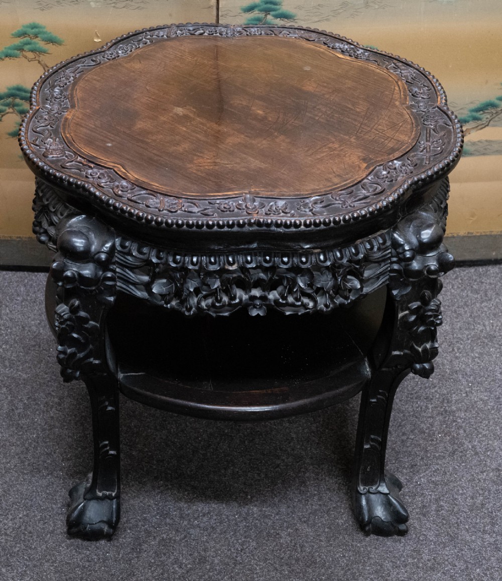 A dark rosewood Chinese occasional table featuring a lobed inset top, deeply carved floral aprons, curved legs, and a lower tier shelf.