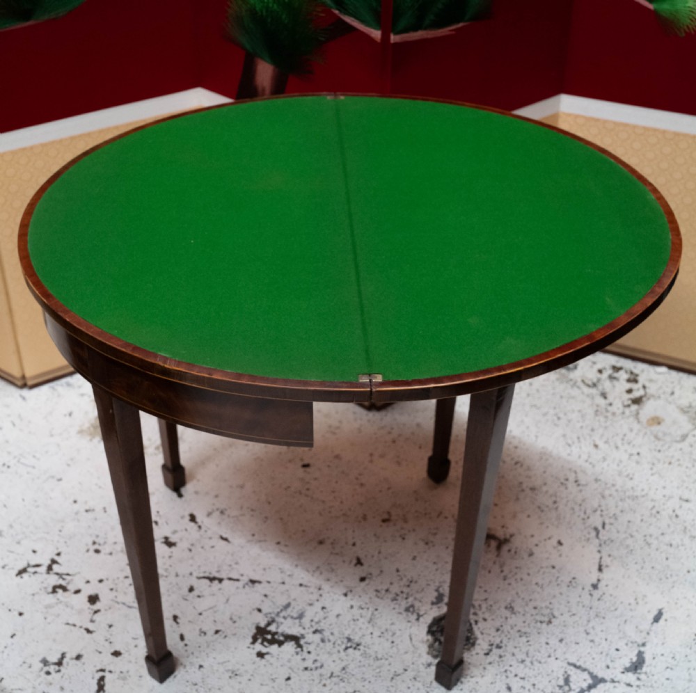 A George III circular mahogany card table with a green baize playing surface and rosewood crossbanding on tapered legs. View IV