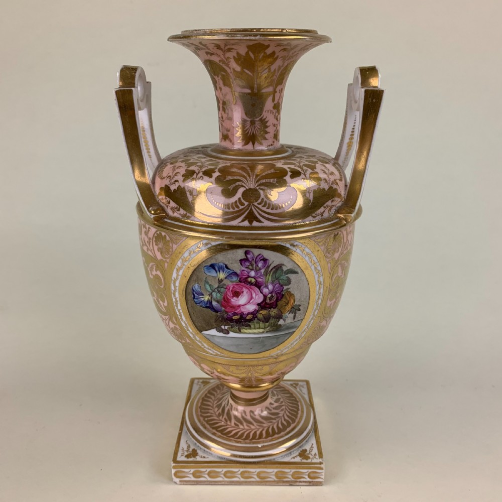 A Regency Derby porcelain urn featuring twin handles, a pink ground with extensive gold scrollwork, and a central hand-painted floral medallion set on a square plinth.