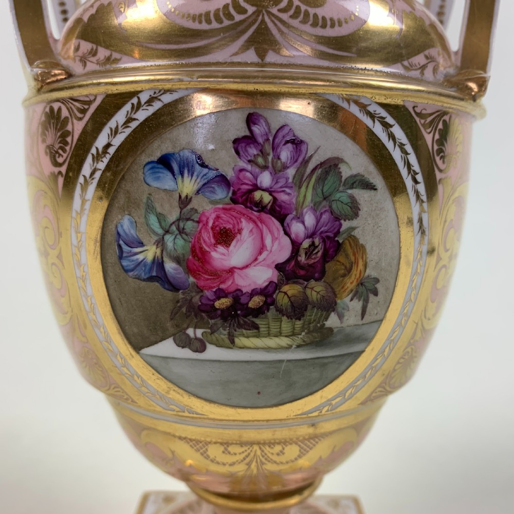 A Regency Derby porcelain urn featuring twin handles, a pink ground with extensive gold scrollwork, and a central hand-painted floral medallion set on a square plinth. View II