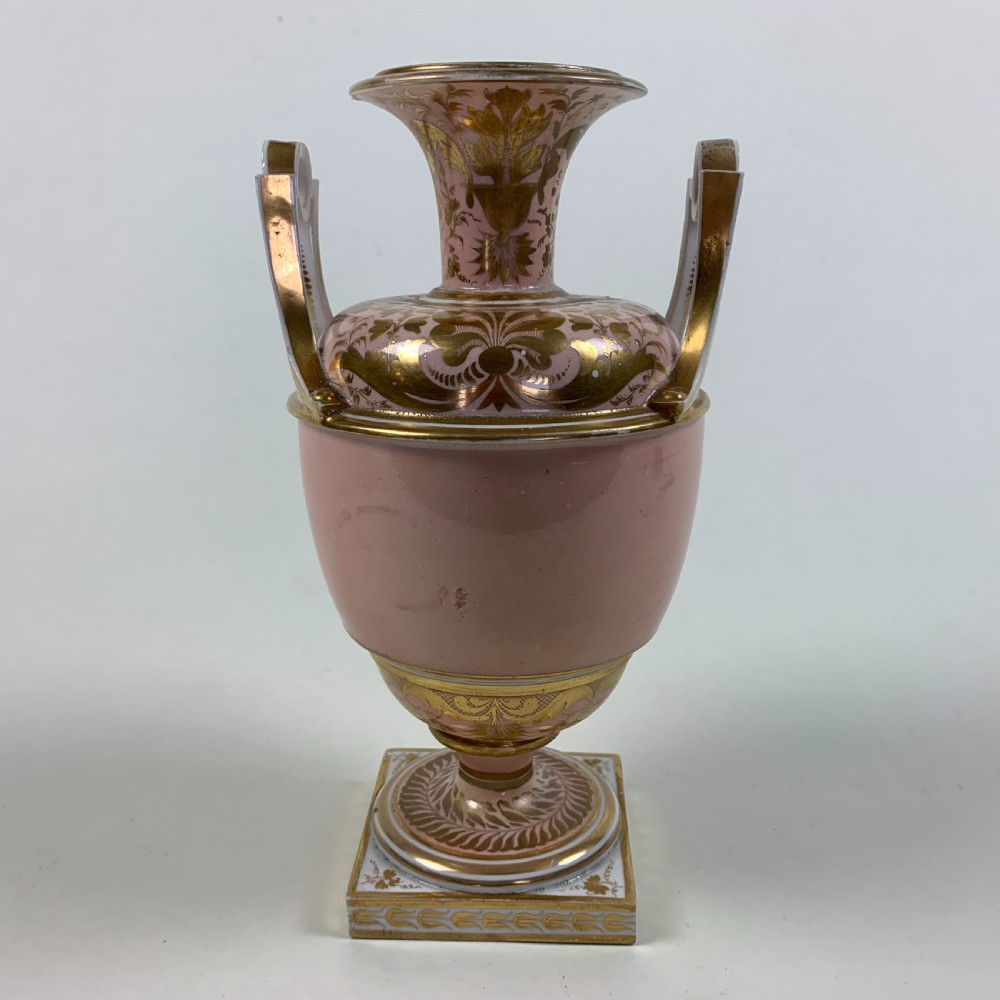 A Regency Derby porcelain urn featuring twin handles, a pink ground with extensive gold scrollwork, and a central hand-painted floral medallion set on a square plinth. View III