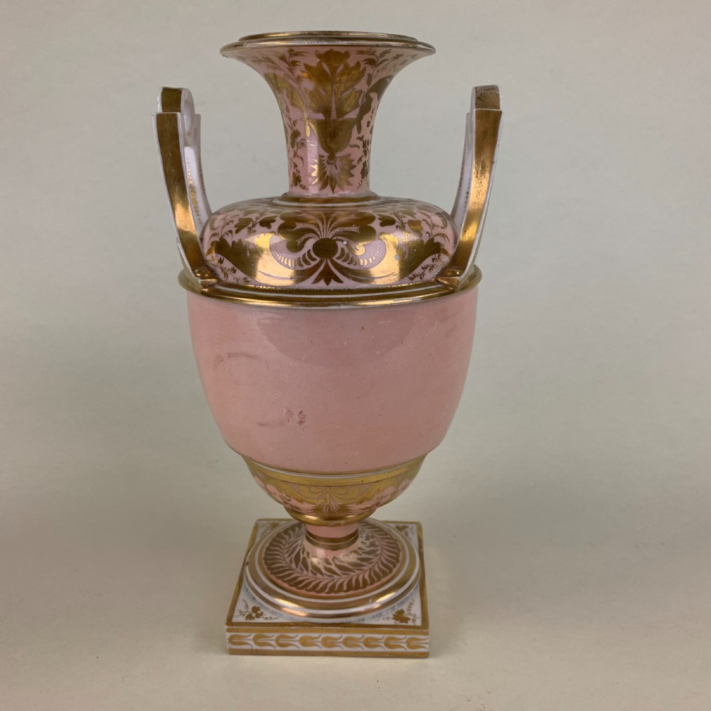 A Regency Derby porcelain urn featuring twin handles, a pink ground with extensive gold scrollwork, and a central hand-painted floral medallion set on a square plinth. View V