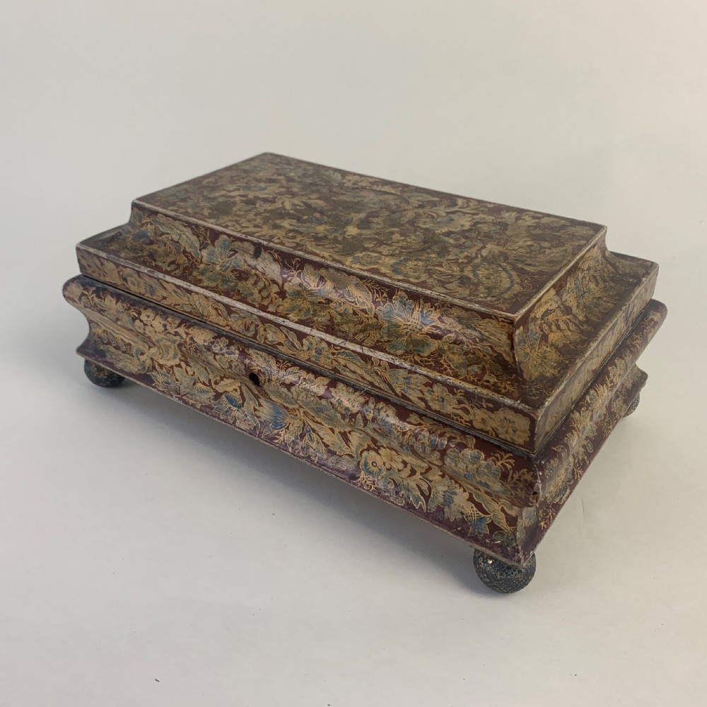 A rectangular early 19th-century Regency wooden games box, hand-painted with intricate botanical patterns in burgundy, gold, and green, featuring a sarcophagus-style lid and small ball feet.