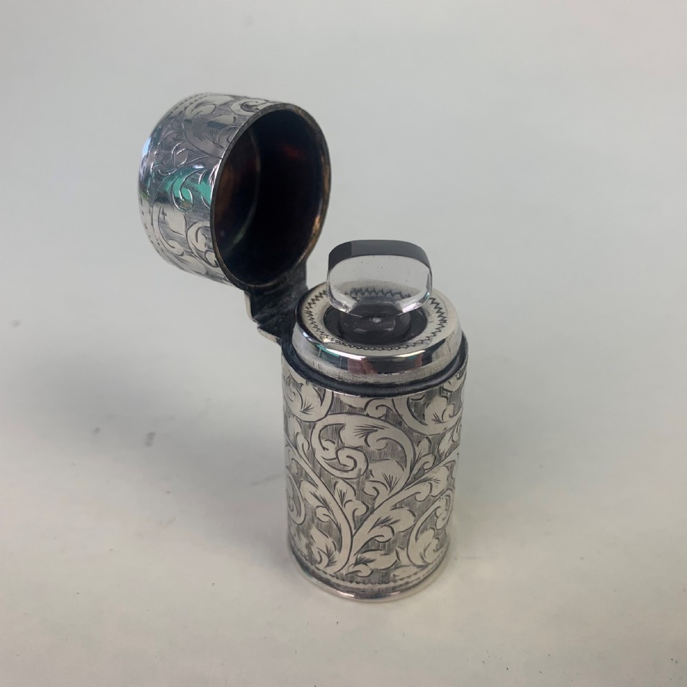 An engraved cylindrical silver scent bottle case with an open hinged lid displaying an internal clear glass bottle with a stopper.