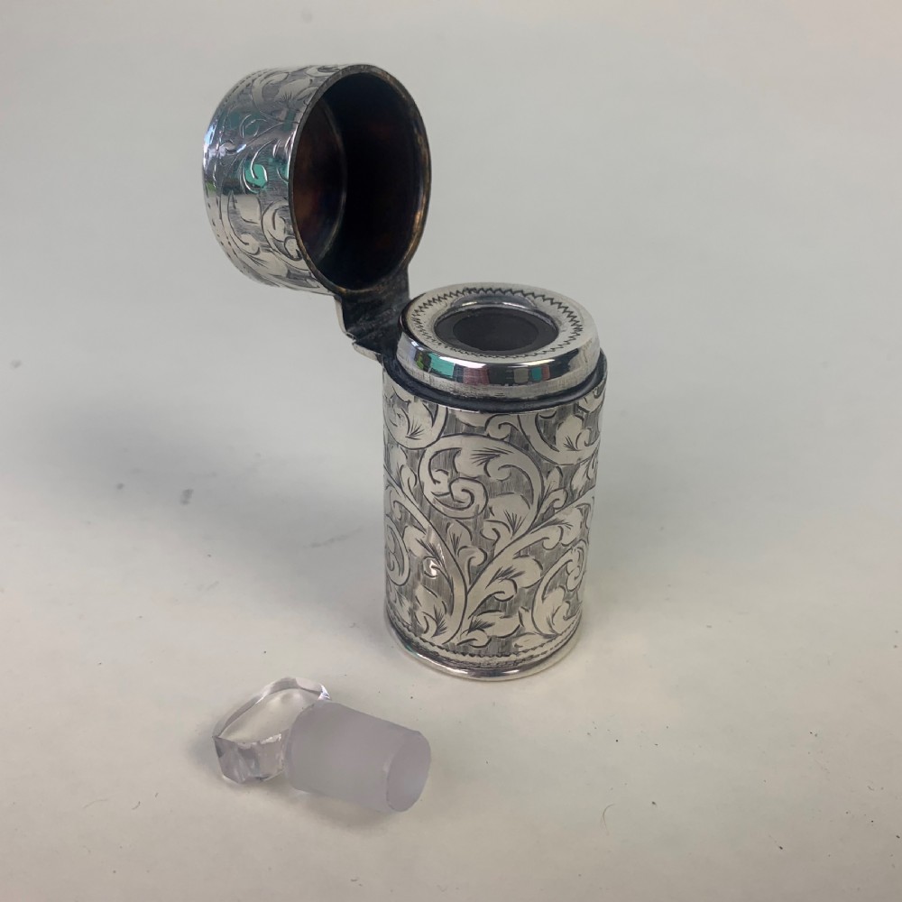 An engraved cylindrical silver scent bottle case with an open hinged lid displaying an internal clear glass bottle with a stopper. View V