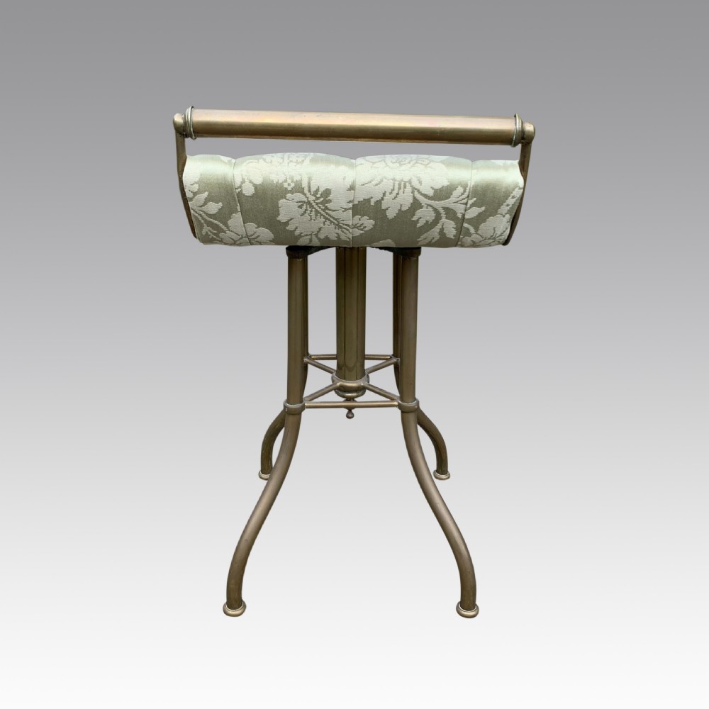 An upright brass pianochello music stool featuring an upholstered cream patterned seat and a curved back rail. View III