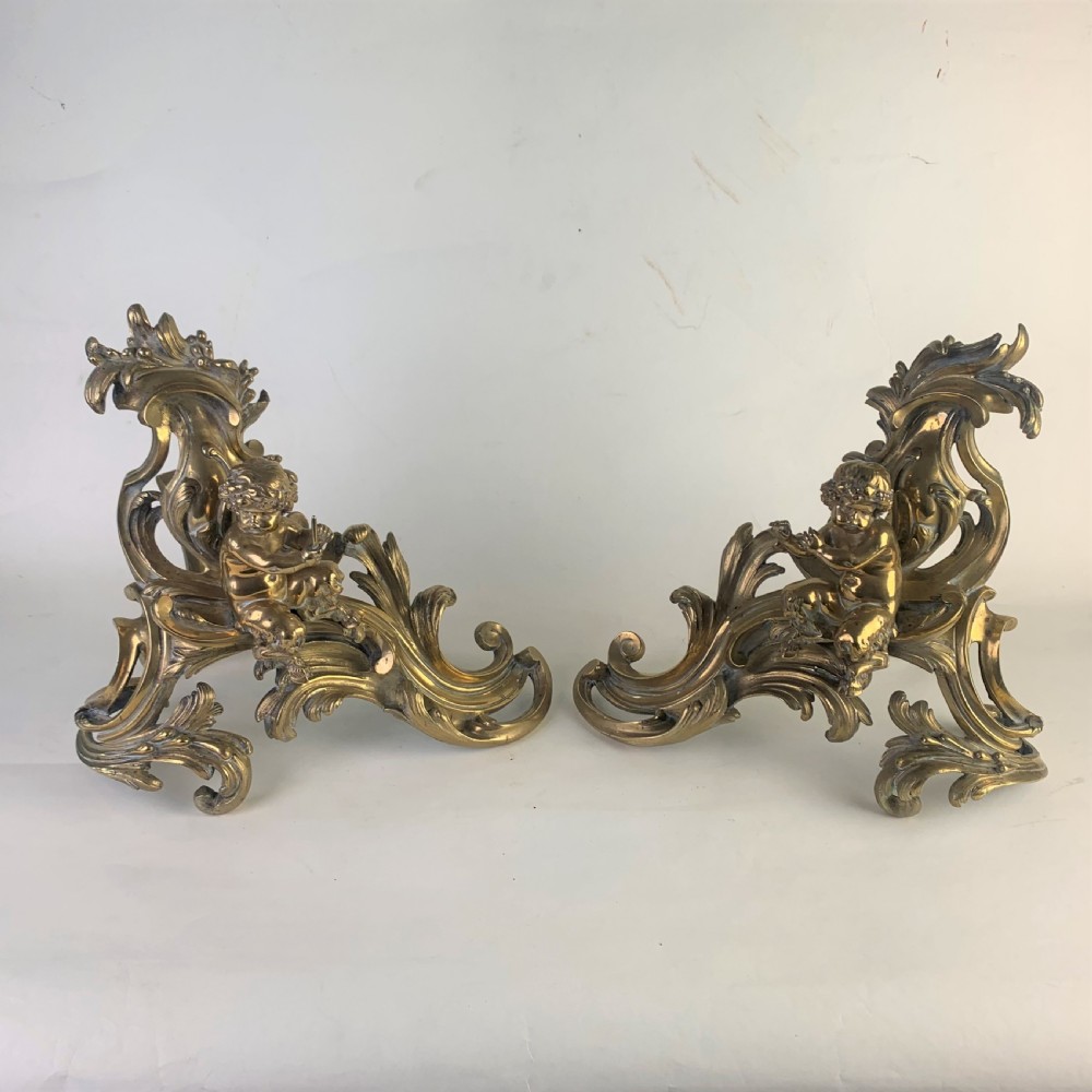 A pair of ornate gilded brass chenets featuring sculpted cherub figures seated amidst asymmetrical leaf scrolls and rocaille motifs.