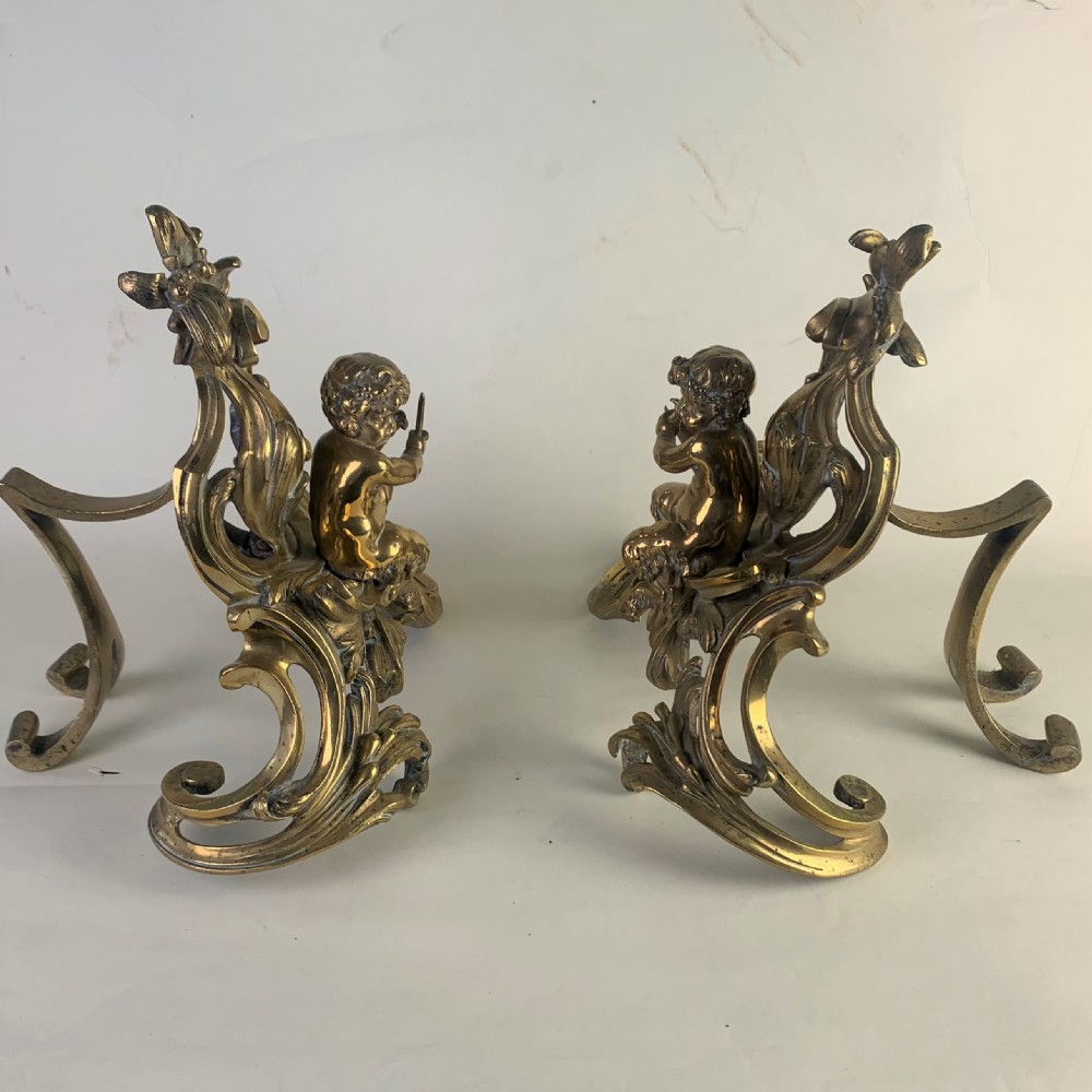 A pair of ornate gilded brass chenets featuring sculpted cherub figures seated amidst asymmetrical leaf scrolls and rocaille motifs. View II