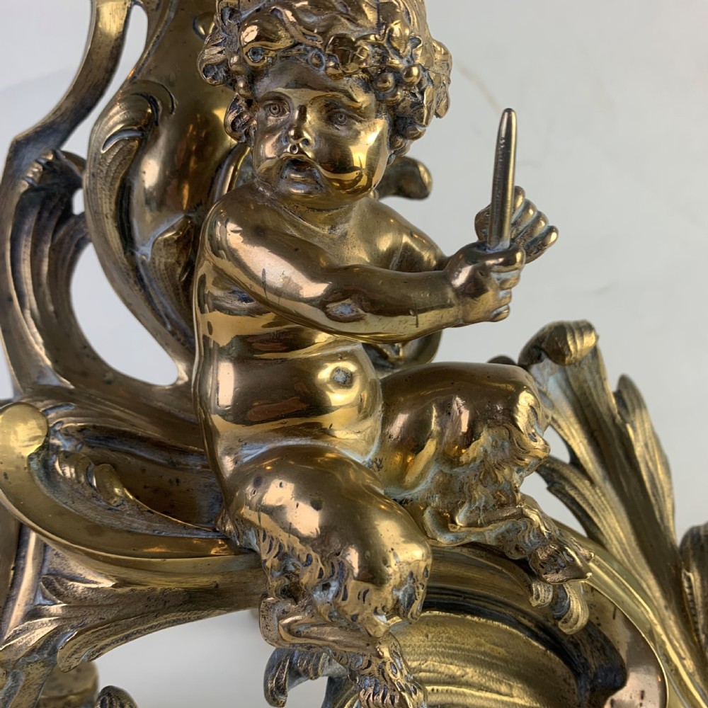 A pair of ornate gilded brass chenets featuring sculpted cherub figures seated amidst asymmetrical leaf scrolls and rocaille motifs. View IV