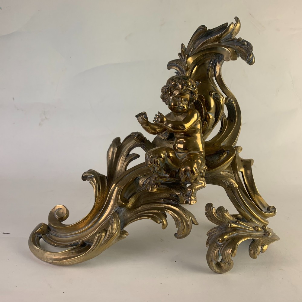 A pair of ornate gilded brass chenets featuring sculpted cherub figures seated amidst asymmetrical leaf scrolls and rocaille motifs. View V