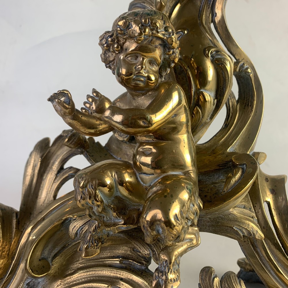 A pair of ornate gilded brass chenets featuring sculpted cherub figures seated amidst asymmetrical leaf scrolls and rocaille motifs. View VII