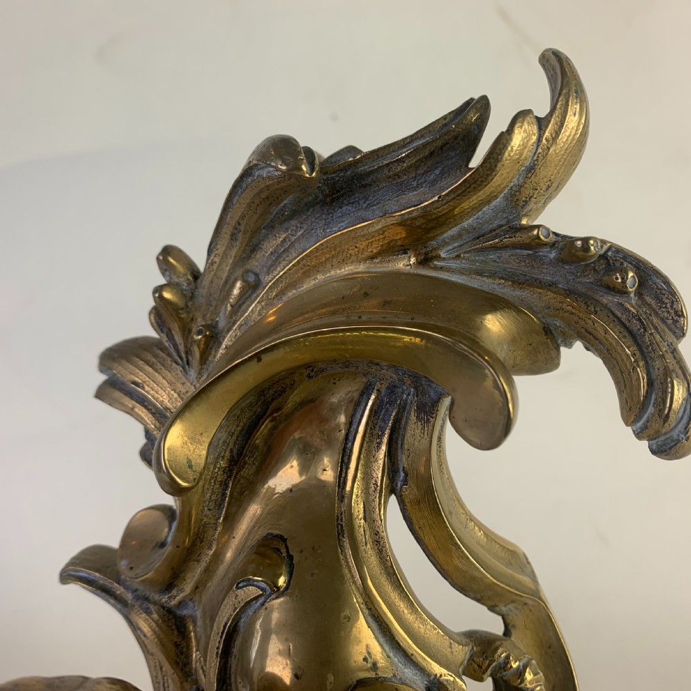 A pair of ornate gilded brass chenets featuring sculpted cherub figures seated amidst asymmetrical leaf scrolls and rocaille motifs. View IX