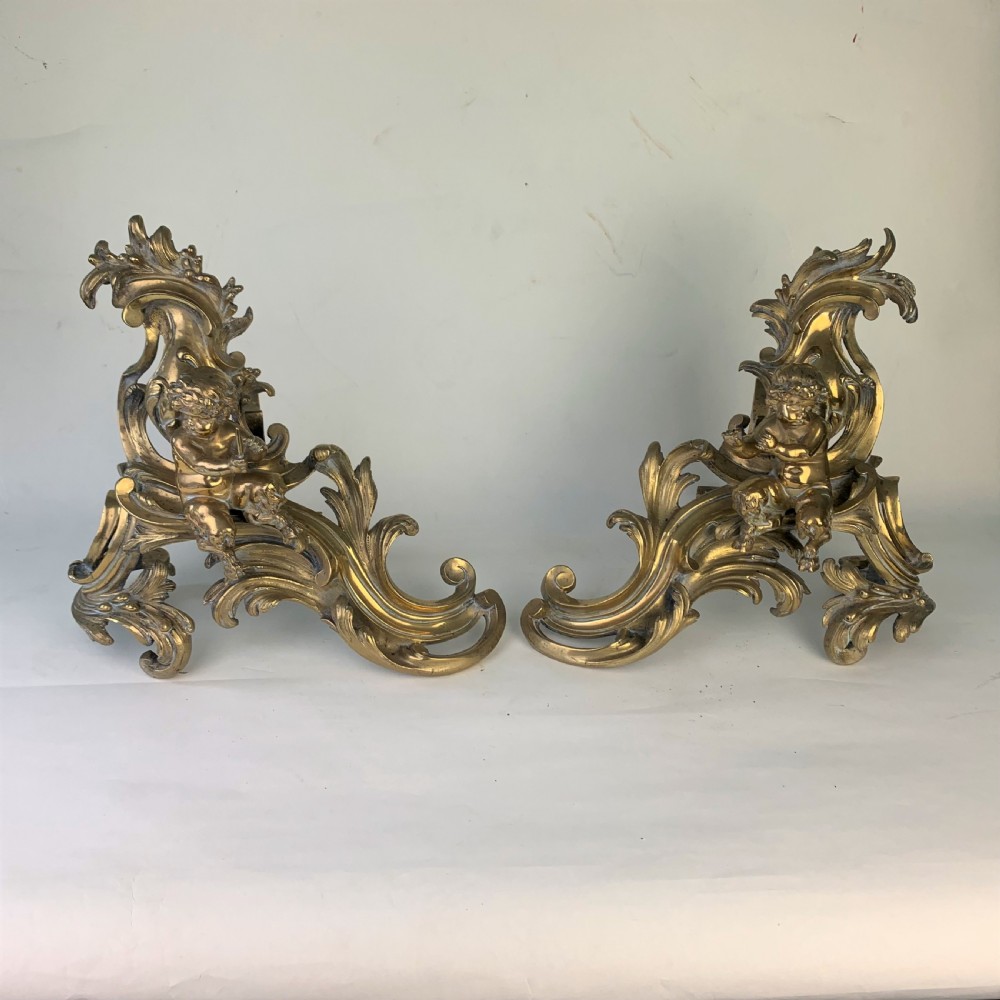 A pair of ornate gilded brass chenets featuring sculpted cherub figures seated amidst asymmetrical leaf scrolls and rocaille motifs. View X