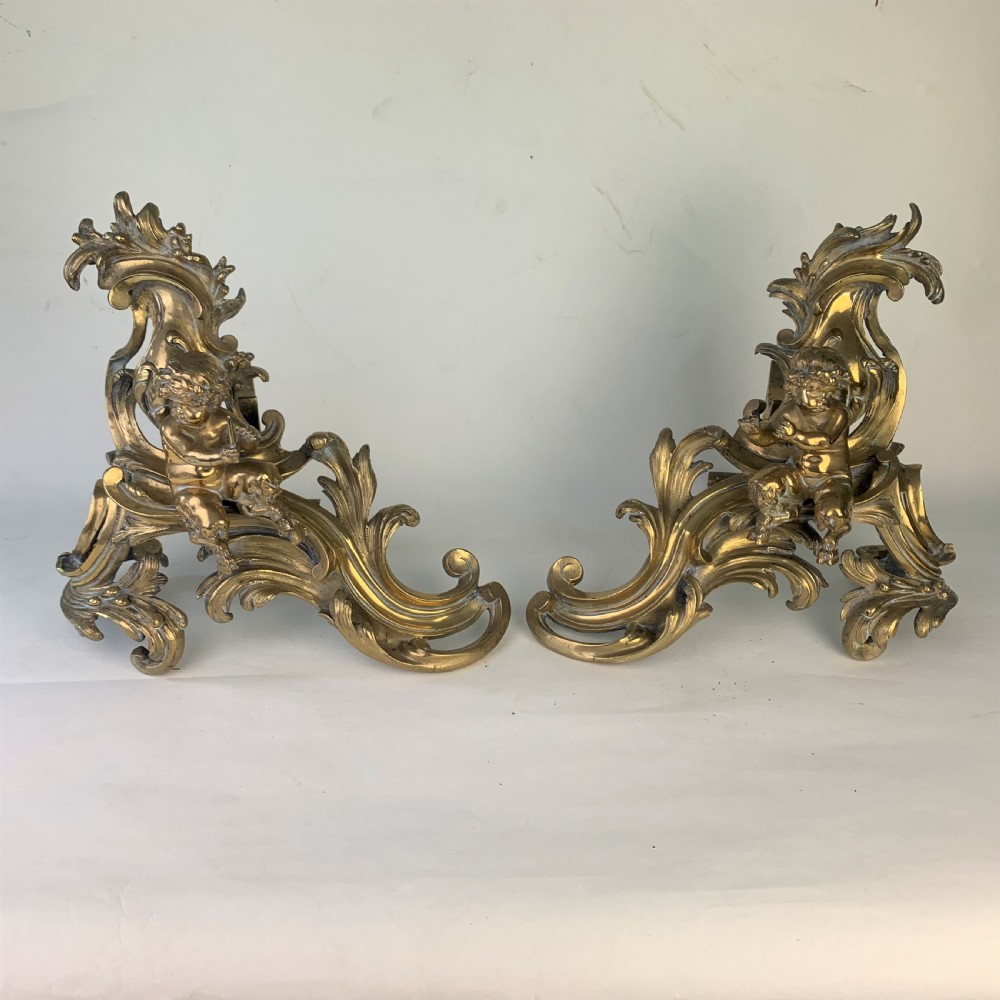 A pair of ornate gilded brass chenets featuring sculpted cherub figures seated amidst asymmetrical leaf scrolls and rocaille motifs. View XI
