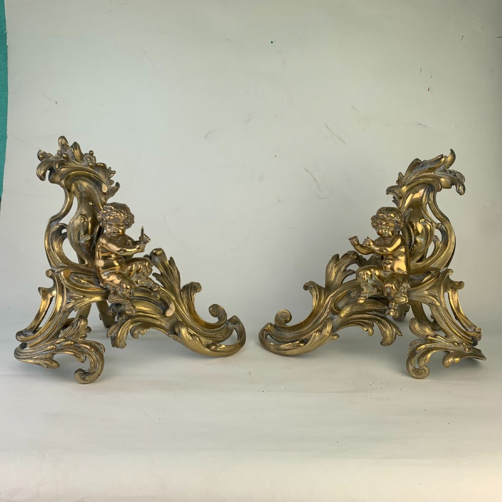A pair of ornate gilded brass chenets featuring sculpted cherub figures seated amidst asymmetrical leaf scrolls and rocaille motifs. View XII