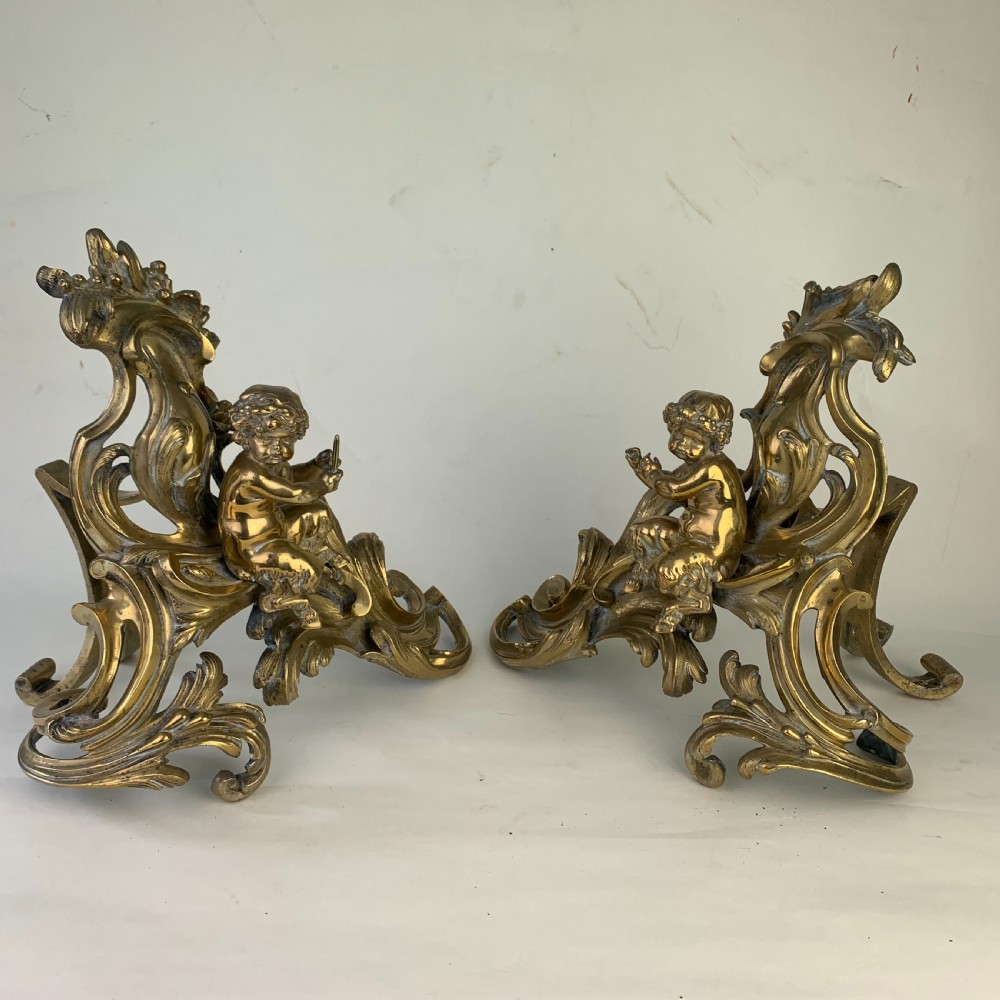 A pair of ornate gilded brass chenets featuring sculpted cherub figures seated amidst asymmetrical leaf scrolls and rocaille motifs. View XIII