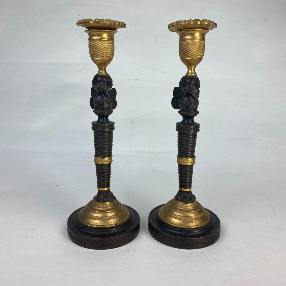 A pair of antique patinated bronze and ormolu candlesticks featuring central putti busts supporting urn sconces set on circular gilded bases.