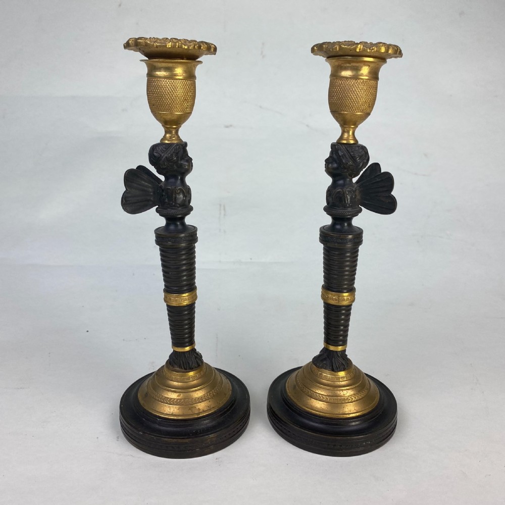 A pair of antique patinated bronze and ormolu candlesticks featuring central putti busts supporting urn sconces set on circular gilded bases. View II