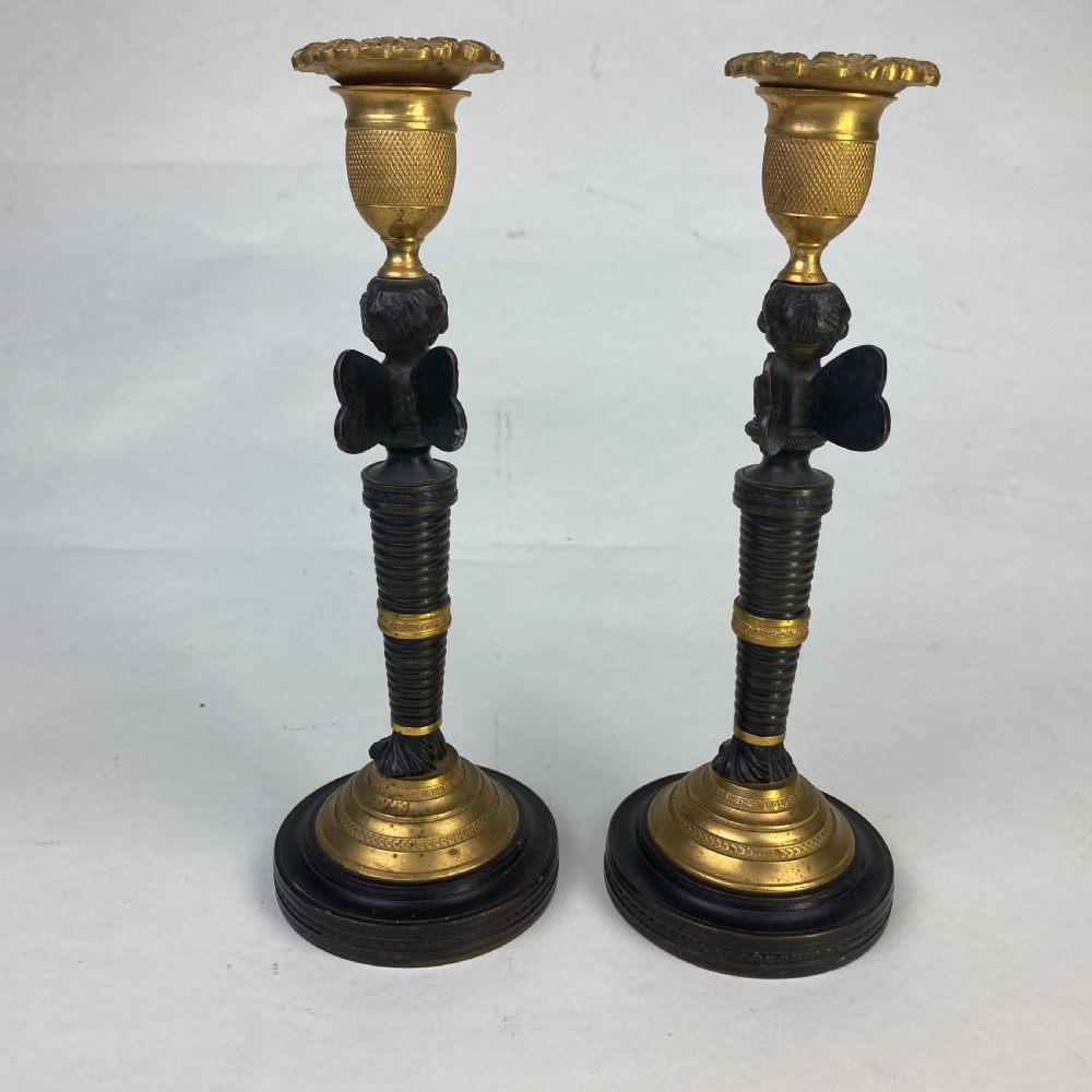 A pair of antique patinated bronze and ormolu candlesticks featuring central putti busts supporting urn sconces set on circular gilded bases. View V