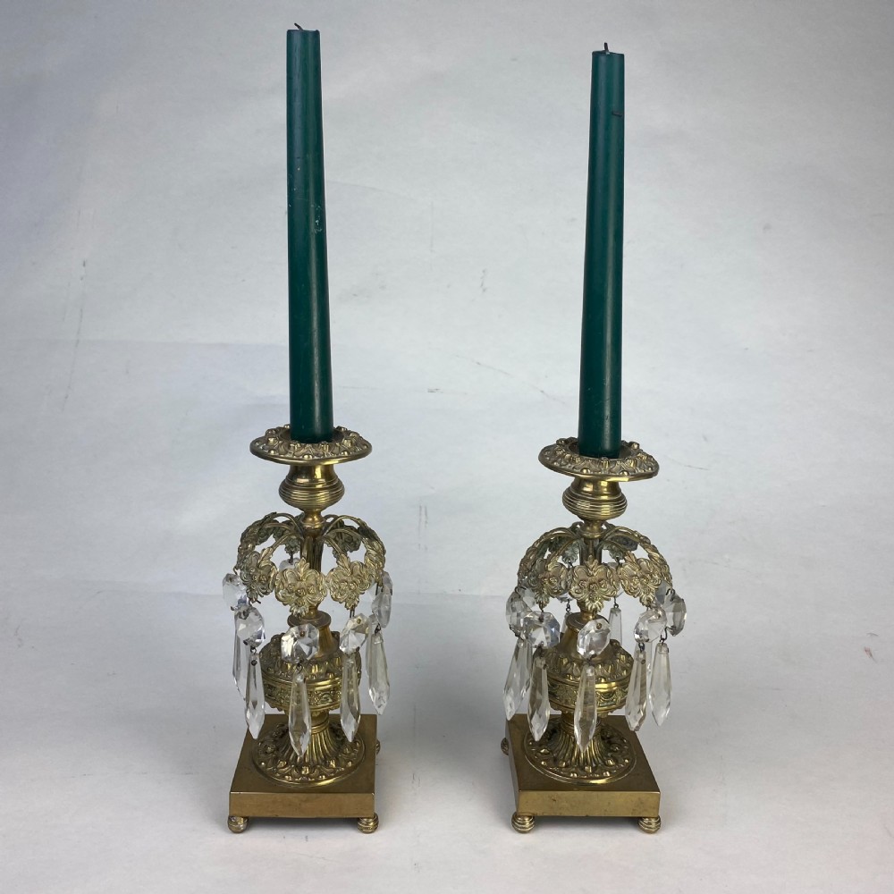 A pair of ornate gilt metal candlesticks featuring square plinth bases, decorative scrollwork rims, and clear cut glass lustre drops hanging around the column.