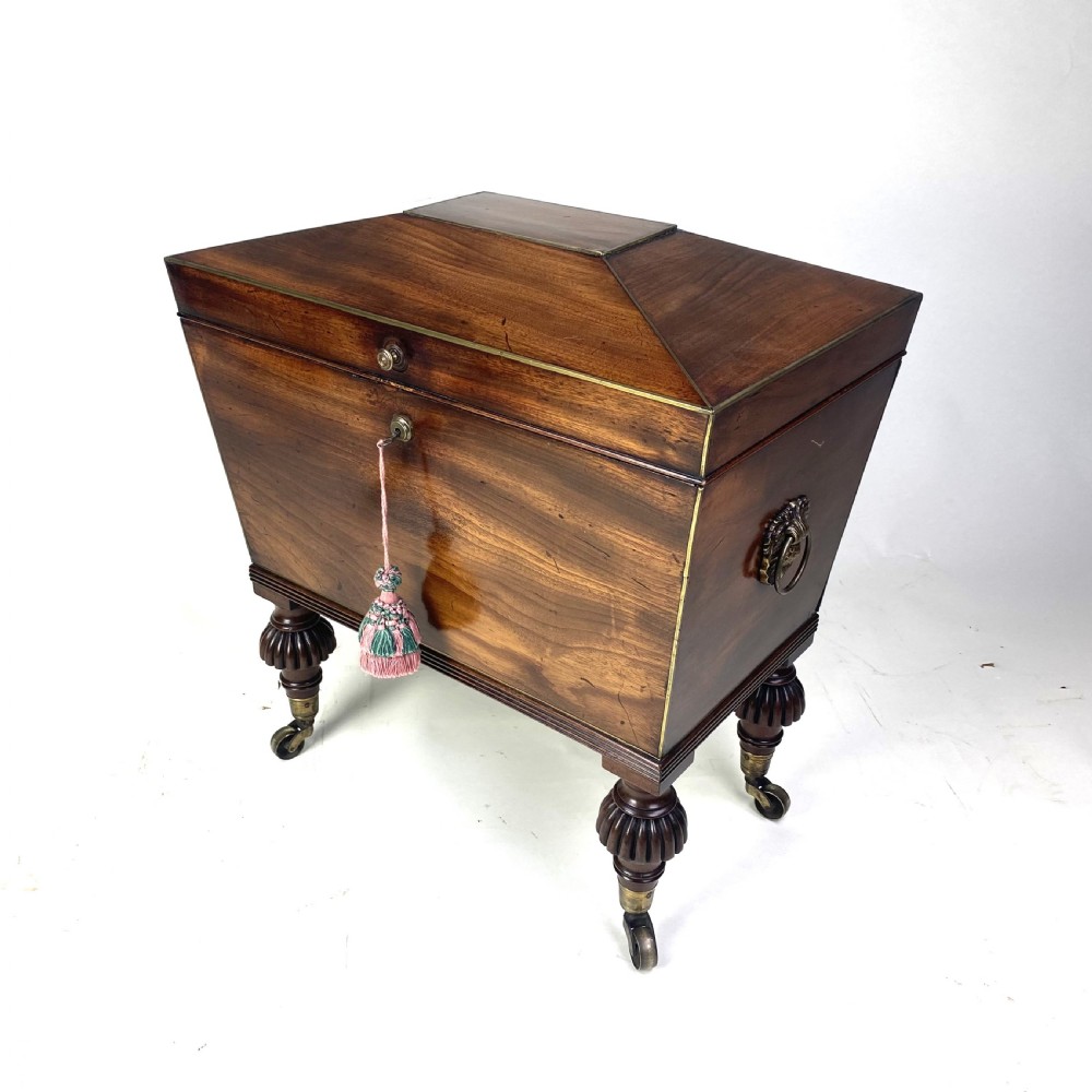 A Regency mahogany sarcophagus-shaped cellarette featuring brass inlay, side lion mask handles, and fluted legs with castors.