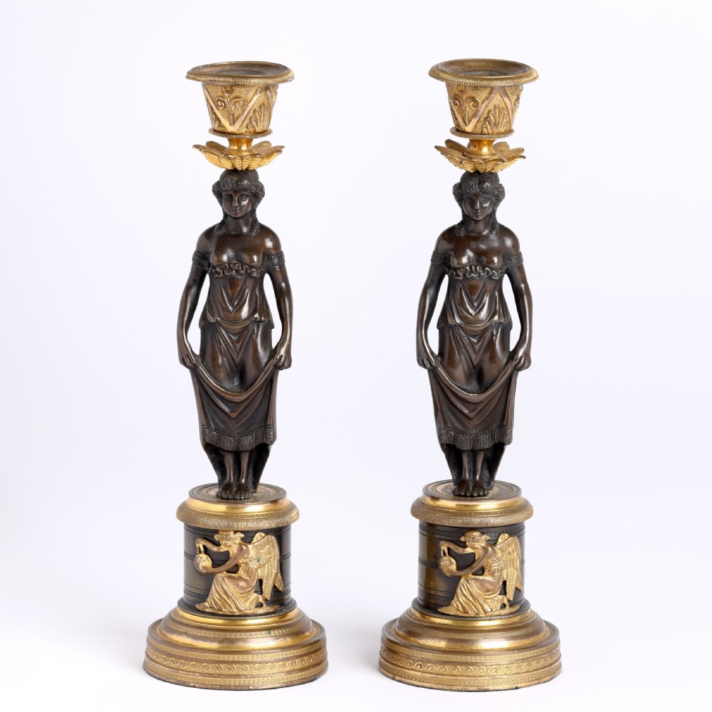 A pair of antique dark bronze figural candlesticks depicting classical draped female figures standing on ornate round gilt bronze bases holding acanthus leaf sconces.