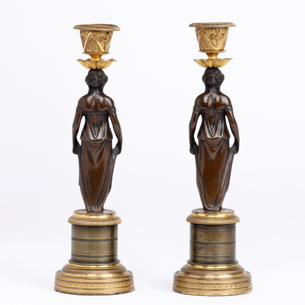 A pair of antique dark bronze figural candlesticks depicting classical draped female figures standing on ornate round gilt bronze bases holding acanthus leaf sconces. View II