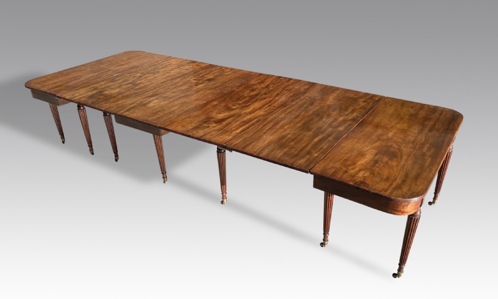An extended mahogany dining table displaying multiple leaf sections, reeded tapered legs, and brass castors. View III