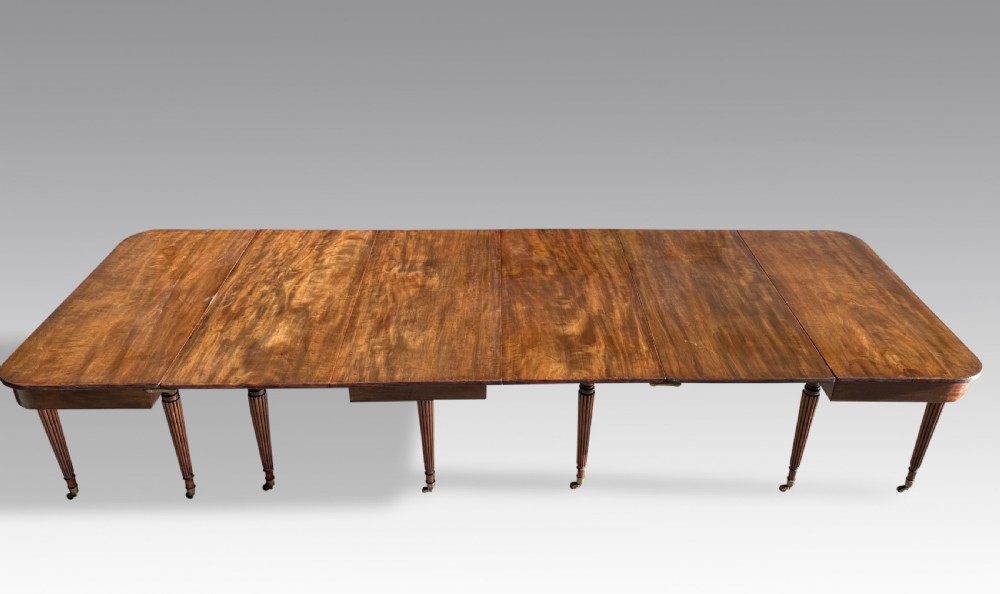 An extended mahogany dining table displaying multiple leaf sections, reeded tapered legs, and brass castors. View IV