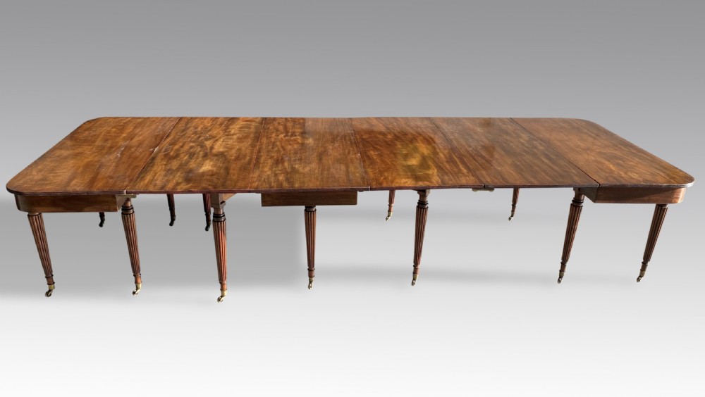 An extended mahogany dining table displaying multiple leaf sections, reeded tapered legs, and brass castors. View V