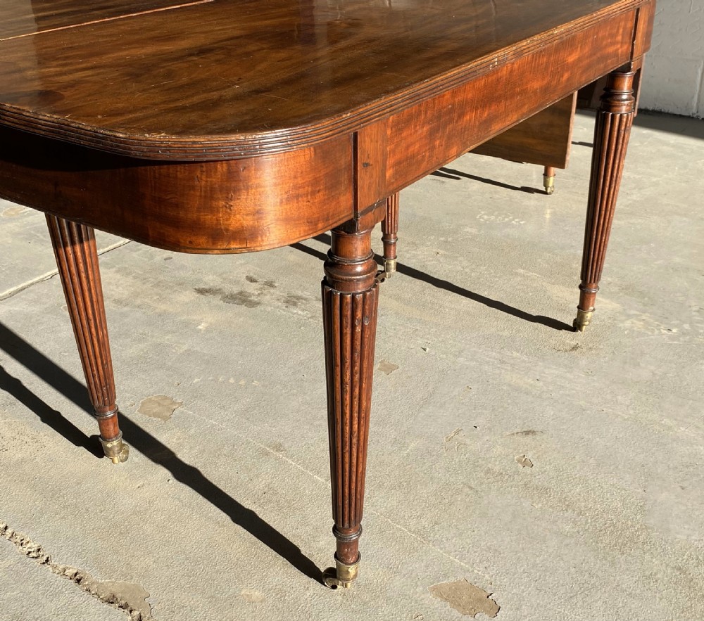 An extended mahogany dining table displaying multiple leaf sections, reeded tapered legs, and brass castors. View VI
