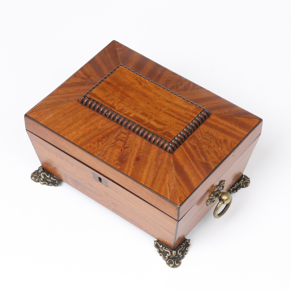 A Regency style satinwood casket box featuring a tapered sarcophagus shape, carved lid trim, brass claw feet, and side ring handles. View V