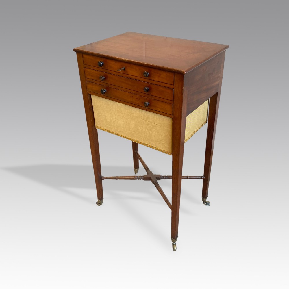 A Georgian mahogany needlework table featuring three drawers, a fabric work pouch, turned X-frame stretcher, and brass castors.