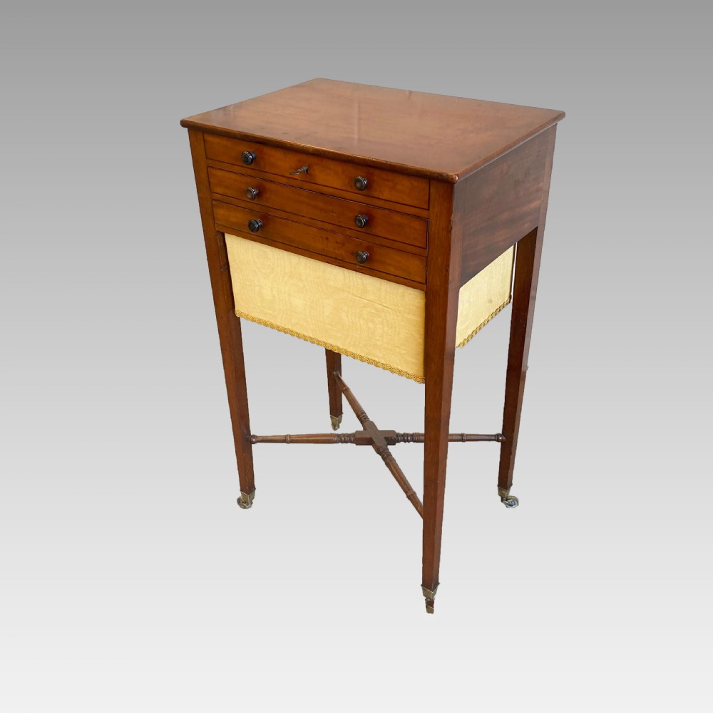 A Georgian mahogany needlework table featuring three drawers, a fabric work pouch, turned X-frame stretcher, and brass castors. View V