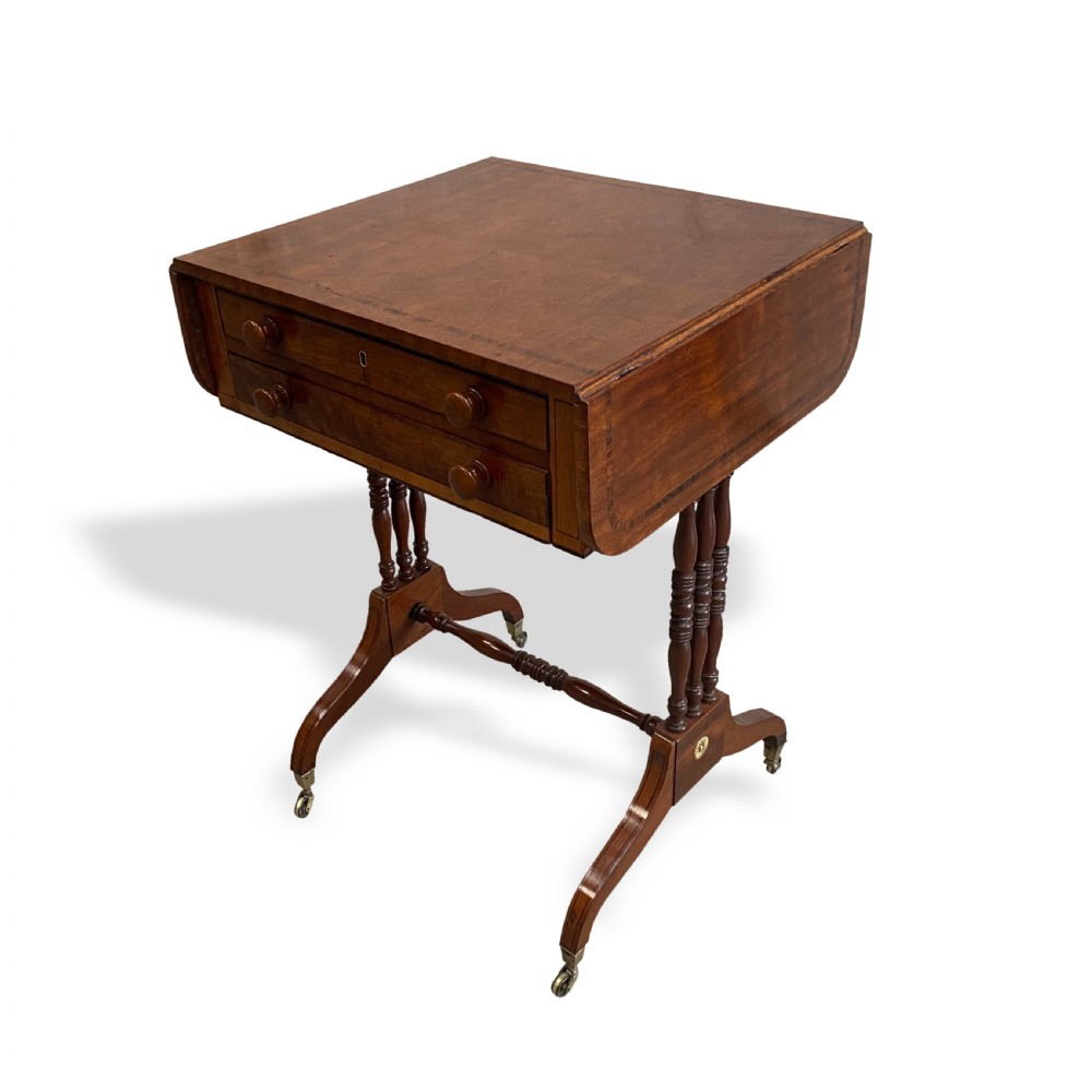 An antique Regency mahogany drop-flap occasional table featuring two drawers with wooden knobs, double turned upright pedestals, and splayed legs on brass castors. View II