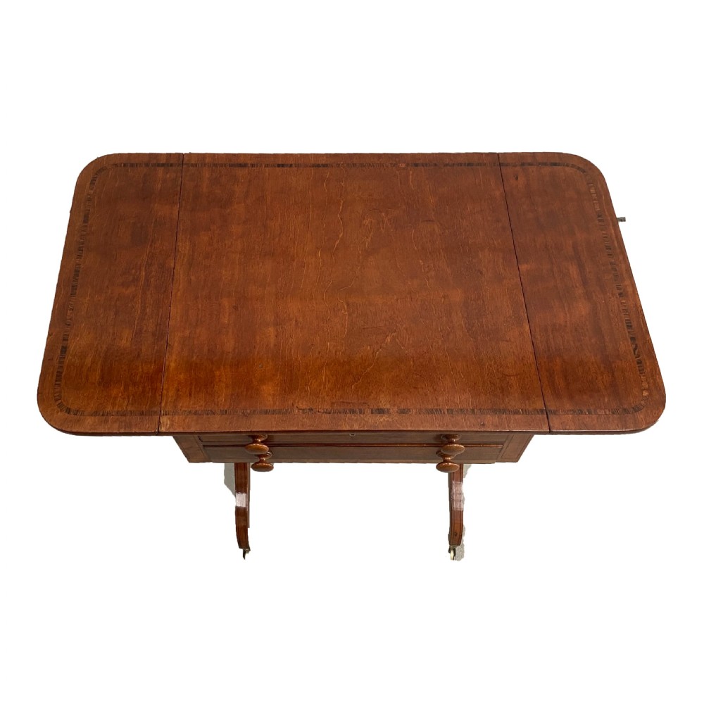 An antique Regency mahogany drop-flap occasional table featuring two drawers with wooden knobs, double turned upright pedestals, and splayed legs on brass castors. View V