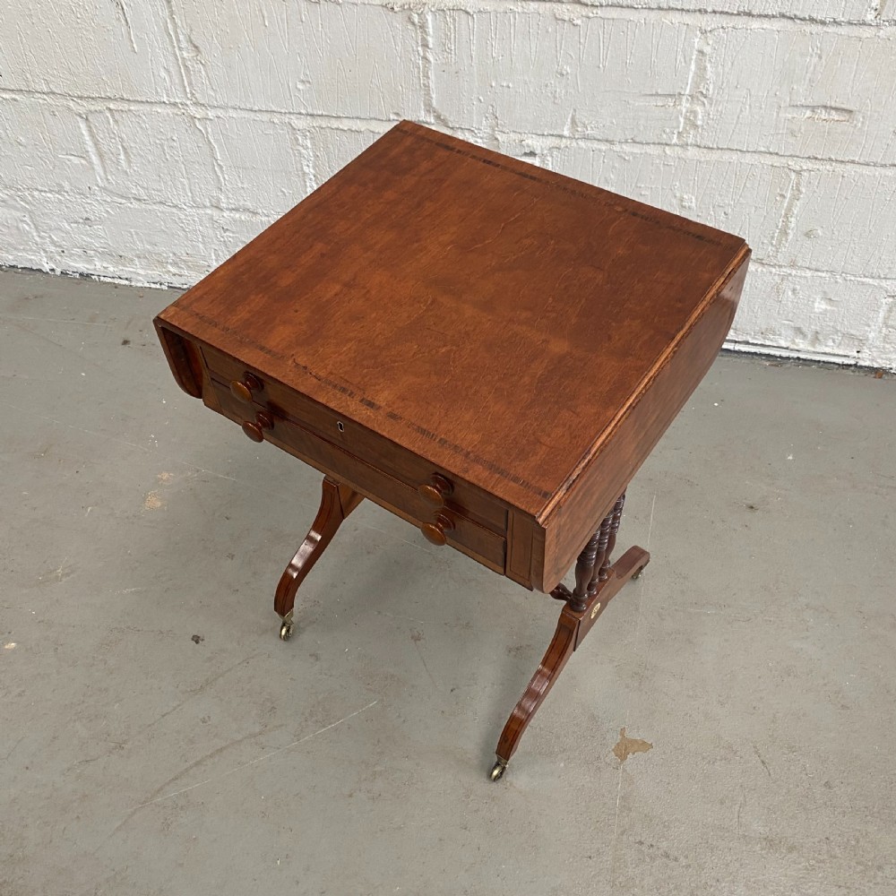 An antique Regency mahogany drop-flap occasional table featuring two drawers with wooden knobs, double turned upright pedestals, and splayed legs on brass castors. View VI