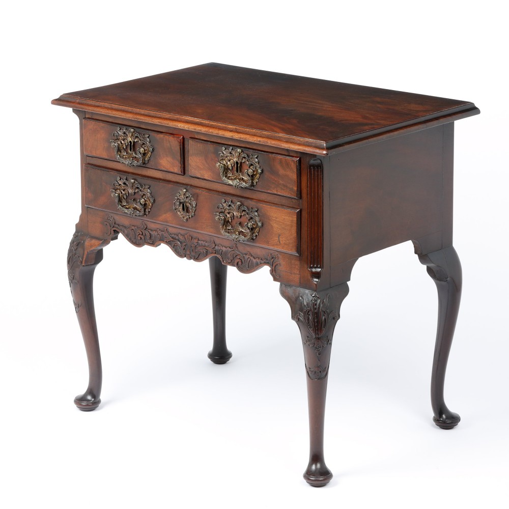 A George II mahogany lowboy side table featuring three drawers, ornate brass backplate handles, carved cabriole legs, and pad feet.