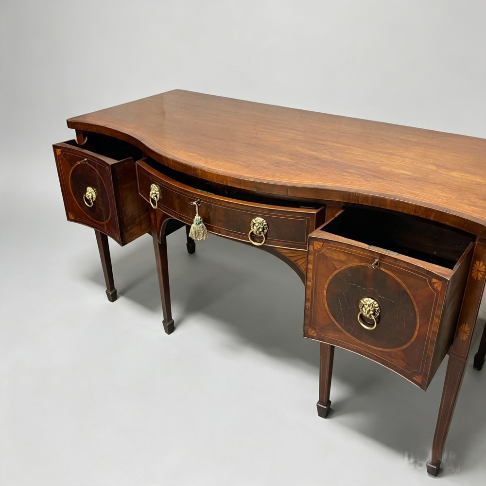 An antique mahogany serpentine-front sideboard featuring decorative wood inlays, brass lion-head ring handles, and square tapered legs. View VII