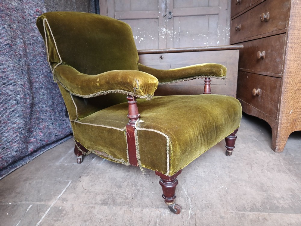 An antique Victorian low easy chair upholstered in olive green velvet with turned wooden arm supports and legs on brass castors.